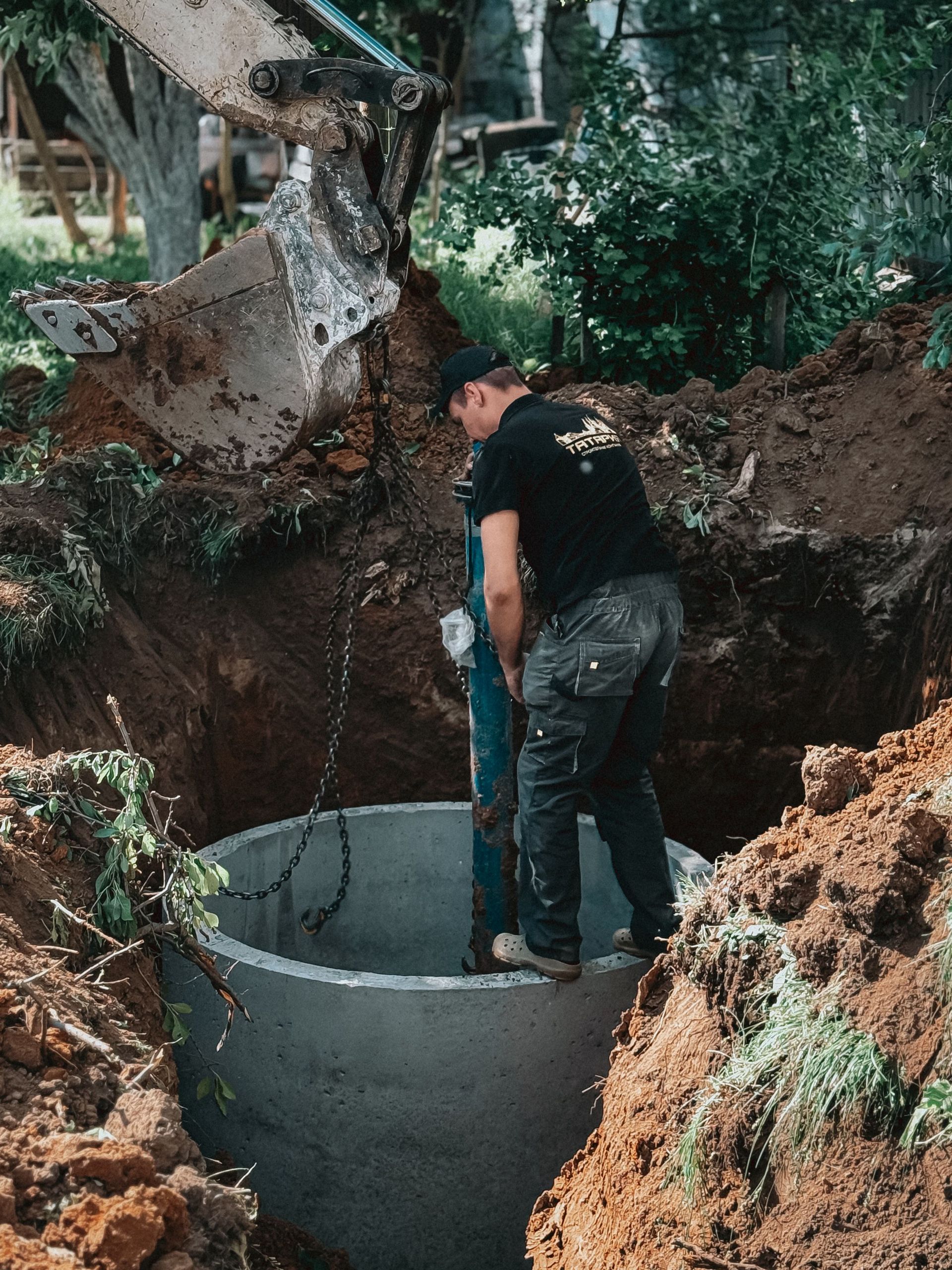 A construction worker operates heavy machinery to install a concrete pipe into the ground, surrounded by dug soil.
